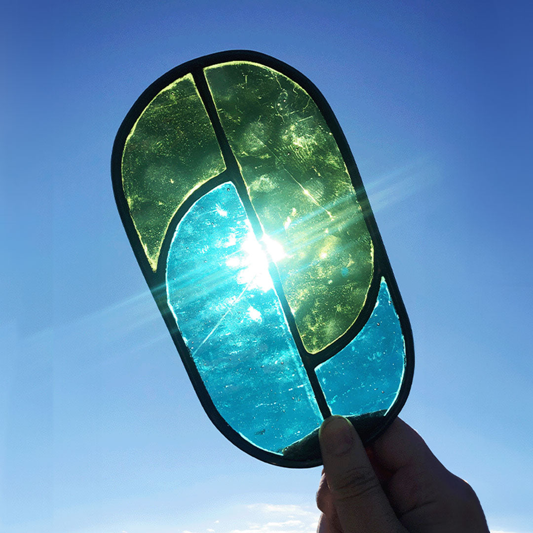 Connected oval in teal and moss green held up to the sun to show the colour of the glass impacted by sunlight. This image shows the glass occlusion (line) in the teal pane